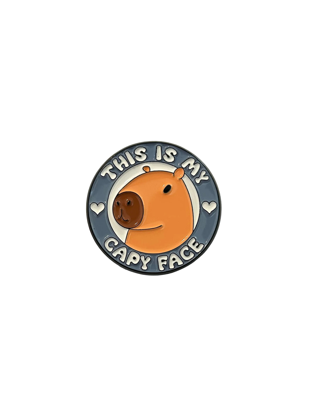 A circular enamel pin featuring a cute orange capybara face with the text 'THIS IS MY CAPY FACE' in white, held by a person's fingers over a white sheet of paper on a wooden surface.